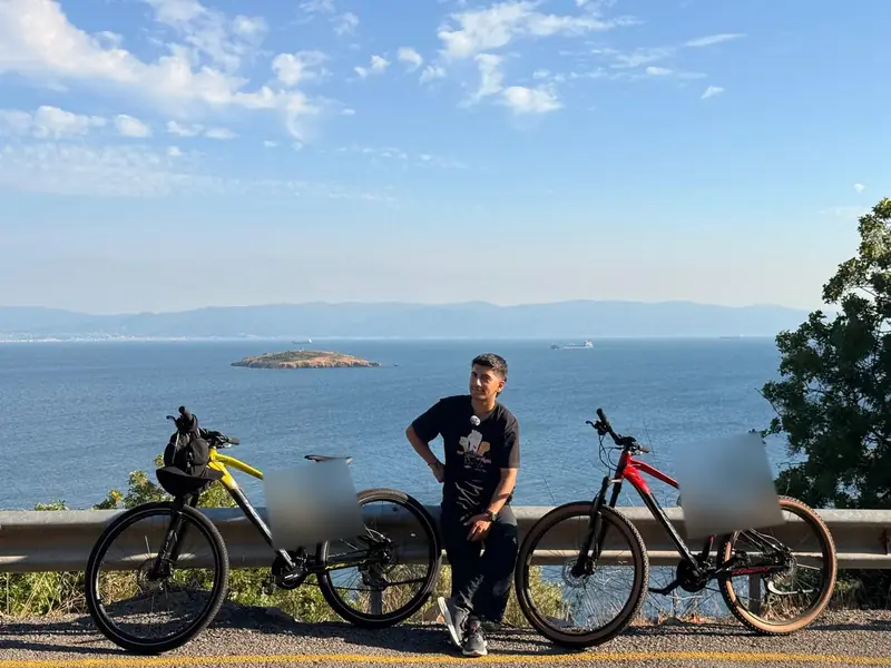 Yiğit, geniş bir kıyıya bakan yolda, iki bisikletle; ada ve dağlar