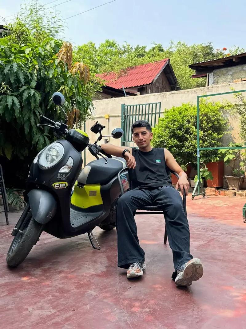 Yiğit, Luang Prabang avlusunda sandalyede, yanında siyah-sarı bir Honda scooter